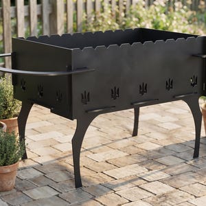 Könnte beinhalten: Ein rechteckiger Holzkohlegrill aus schwarzem Metall mit gezackter Oberkante und ausgestanzten Dreizacksymbolen an den Seitenwänden. Der Grill steht auf vier geschwungenen Beinen und verfügt über zwei halbkreisförmige Seitengriffe.