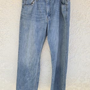 May include: A pair of light blue denim jeans featuring a classic five-pocket design, a button fly closure, and a faded wash finish. The jeans are constructed with visible stitching along the seams and are displayed hanging on a wire hanger.