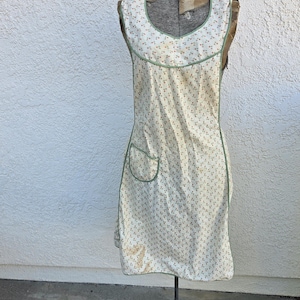 May include: A sleeveless apron featuring a cream-colored fabric with a small-scale floral pattern in shades of red and green. The apron has a rounded neckline, green piping trim along the edges, and a single patch pocket on the right side.