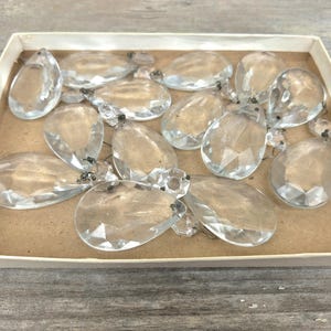 Op de afbeelding: Een verzameling heldere, facetgeslepen kristallen kroonluchterhangers in een kartonnen doos. De hangers zijn druppelvormig en hebben kleine metalen bevestigingen. De doos heeft een lichtbeige kleur.