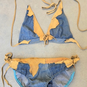 May include: A two-piece bikini set crafted from blue denim fabric and tan leather trim. The triangle top features leather edging and long leather tie straps. The matching bikini bottom includes leather waistband detailing and side tie closures with blue fabric lining.