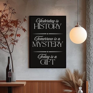 Könnte beinhalten: Ein schwarzer, gerahmter Kunstdruck mit dem weißen Schriftzug "Yesterday is HISTORY, Tomorrow is a MYSTERY, Today is a GIFT". Der Druck hängt in einem Raum mit neutralen Farben, einer Kugelleuchte und einem Holztisch.