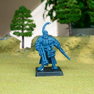 May include: A blue miniature figurine of a historical soldier, standing on a black base. The figure is detailed with a feathered hat, ornate clothing, and two rifles. The background features miniature trees and a grassy terrain.