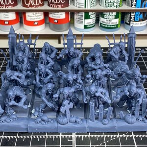 Puede incluir: Un grupo de figuras de zombis en miniatura de color azul grisáceo, con lápidas, calabazas y un esqueleto. Las figuras están sobre una base rectangular, lo que sugiere un juego de mesa o un diorama.