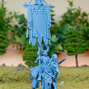 Peut inclure: Miniature bleu clair d'un chevalier à cheval, tenant une bannière. Le chevalier est en armure et porte une épée. La bannière est ornée d'éléments décoratifs. La miniature est placée sur un fond de feuillage vert et une surface herbeuse.