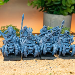 May include: A group of blue fantasy miniatures, each mounted on a spider-like creature. The figures are detailed with weapons and armor, set on black bases. The background includes greenery and a neutral-toned surface.
