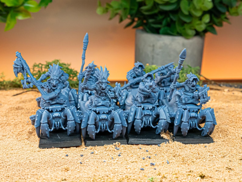 May include: A group of blue fantasy miniatures, each mounted on a spider-like creature. The figures are detailed with weapons and armor, set on black bases. The background includes greenery and a neutral-toned surface.