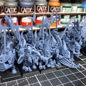 May include: A group of grey miniature fantasy figures, each with distinct features and weapons. The figures are on black bases, with various poses and details, including axes and nets. Paint bottles are visible in the background.