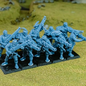 Puede incluir: Un grupo de figuras en miniatura azules de soldados, cada uno con un rifle, están dispuestas sobre una base rectangular negra. Las figuras, con uniformes detallados, se encuentran sobre un fondo verde y herboso.