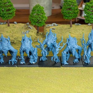 Puede incluir: Cinco figuras de fantasía en miniatura azules, posiblemente para juegos de mesa, están dispuestas sobre una superficie verde y marrón. Cada figura tiene una forma humanoide con una textura de árbol y está montada sobre una base negra. Hay árboles en miniatura en el fondo.