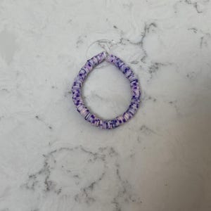 May include: A bracelet made of small, flat, round beads in shades of purple, white, and pink. The beads are strung together to form a circular shape. The bracelet is shown on a white marble surface.