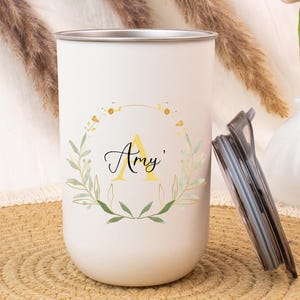 Puede incluir: Vaso blanco con borde plateado, con un diseño de corona floral dorada y verde. El nombre "Amy" está escrito en cursiva, con una letra "A" amarilla en el centro. Una tapa negra descansa junto al vaso. Un pequeño jarrón blanco con tulipanes rosas está en el fondo.