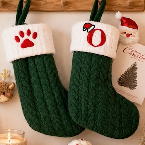 May include: Two dark green cable-knit Christmas stockings with white ribbed cuffs. The left stocking features a red embroidered paw print, while the right stocking displays a red embroidered letter O topped with a small Santa hat graphic.