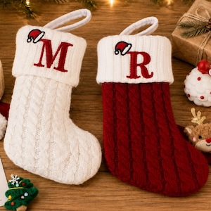 May include: Two cable-knit Christmas stockings, one cream and one deep red, each featuring a white cuff with a red embroidered letter M or R and a small Santa hat icon. The stockings include a white hanging loop and a textured cable pattern.