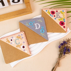 Peut inclure: Ensemble de trois marque-pages d'angle faits à la main avec des motifs floraux brodés et des lettres initiales. Chaque marque-page présente une base en papier brun avec un coin brodé coloré, avec les lettres "C", "D" et "K".