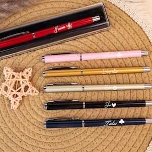 May include: A collection of personalized pens in various colors, including red, pink, gold, silver, black, and navy blue. Each pen has a name or word engraved on it, with decorative symbols. The red pen is in a clear case.