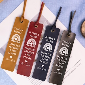 May include: Four leather bookmarks in shades of brown, red, navy, and charcoal. Each bookmark features the text "IT TAKES A VILLAGE" and "THANK YOU FOR BEING A PART OF OURS." with a rainbow graphic and a heart. A leather cord is attached.