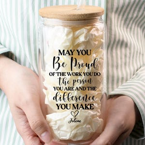 May include: A clear glass tumbler with a bamboo lid and straw, filled with white paper. The tumbler has the text "MAY YOU Be Proud OF THE WORK YOU DO the person YOU ARE AND THE difference YOU MAKE Jolene" in black script.