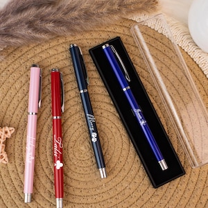 May include: A collection of personalized pens in pink, red, and navy blue, with silver accents. The pens are engraved with names and a clover or paw print design. One blue pen is in a black case with a clear lid.