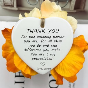 Puede incluir: Un adorno de cerámica blanco en forma de corazón con las palabras "THANK YOU" y un mensaje sincero en texto negro. El adorno está rodeado de vibrantes flores artificiales naranjas y amarillas. Se adjunta una cuerda de yute para colgar.