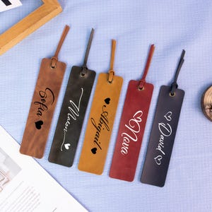 May include: Five leather bookmarks in shades of brown, black, and red, each with a name in white script and a heart symbol. Each bookmark has a matching leather strap attached at the top.