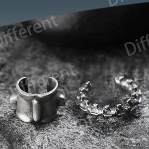 May include: Two silver-toned ear cuffs on a textured gray surface. One cuff has a geometric design, while the other is a simple, open-ended band. The word "Different" is visible in the background.