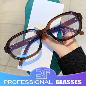 Puede incluir: Gafas con montura marrón y lentes transparentes. Las gafas tienen forma rectangular redondeada y se sostienen sobre un fondo blanco. El texto "PROFESSIONAL GLASSES" es visible en la parte inferior de la imagen.