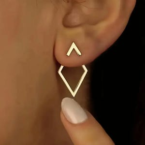 Peut inclure: Boucles d'oreilles géométriques dorées. Les boucles d'oreilles présentent un petit clou triangulaire et une goutte en forme de losange plus grande. Le design est minimaliste et moderne, avec une finition polie. Les boucles d'oreilles sont portées sur une oreille.