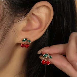 Puede incluir: Pendientes de botón con forma de cereza. Cada pendiente presenta dos cerezas rojas con forma de gema y hojas verdes. Los pendientes se llevan en la oreja, y uno se sostiene en la mano. Los pendientes tienen un engaste de metal dorado.