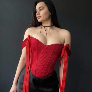 May include: A red corset with off-the-shoulder design and vertical seams. The corset features red ribbon ties at the shoulders. The model is wearing a black choker and a black skirt. The background is a dark gray.