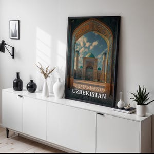 May include: A framed print of a building in Samarkand, Uzbekistan, with a large archway and turquoise domes. The print is displayed on a white cabinet with black vases and a small plant.