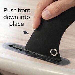 May include: A black surfboard fin being inserted into a slot on a wooden surface. The fin has a white circular design and the text "Push front down into place".