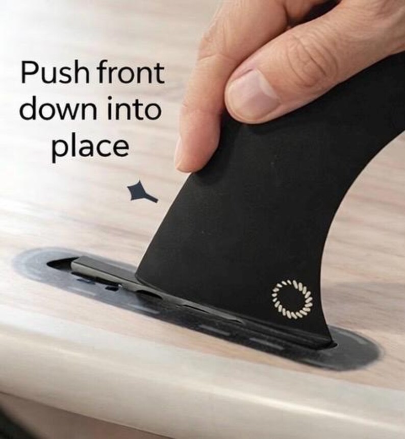 May include: A black surfboard fin being inserted into a slot on a wooden surface. The fin has a white circular design and the text "Push front down into place".