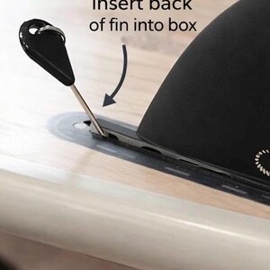 May include: A black fin with a metal pin is being inserted into a slot on a light-colored surface. The text "Insert back of fin into box" is above the fin, with an arrow indicating the direction of insertion. The fin has a curved shape.