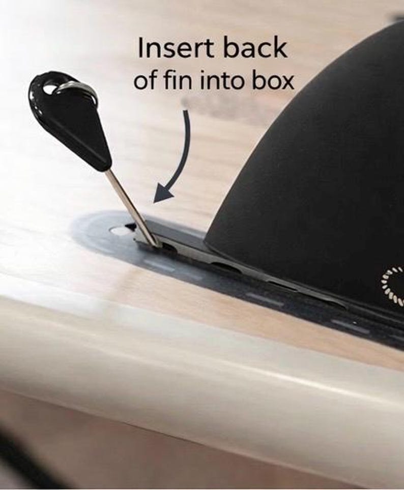 May include: A black fin with a metal pin is being inserted into a slot on a light-colored surface. The text "Insert back of fin into box" is above the fin, with an arrow indicating the direction of insertion. The fin has a curved shape.