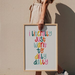May include: A framed print with the text "i literally just want to dilly dally" in colorful, playful lettering. The frame is light-colored wood. The image is held by a person wearing a beige dress and white sneakers.