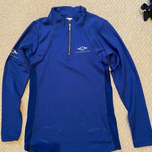 May include: A royal blue equestrian shirt with long sleeves, a gold zip, and the text "LUXE EQUESTRIAN" on the chest and sleeve. The shirt features a mock neck and a fitted design. The equestrian shirt is made of a lightweight, breathable material.