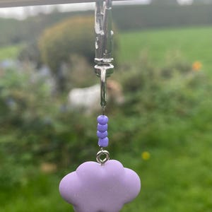 Puede incluir: Un llavero plateado con un dije de nube morada. La nube es de color lavanda suave y está conectada al llavero con un pequeño anillo plateado y una cadena de cuentas moradas. El fondo es una escena exterior verde borrosa.