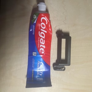 May include: A 6.0 ounce tube of Colgate Cavity Protection fluoride toothpaste with a red, white, and blue label, positioned next to a black plastic toothpaste tube squeezer tool with a rectangular frame and a small winding handle.