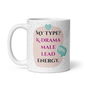 May include: White ceramic mug with the text "MY TYPE? K-DRAMA MALE LEAD ENERGY." in black and purple lettering. A light pink shape with a teal heart and a speech bubble that says "OPPA!" are also on the mug. The mug has a white handle.
