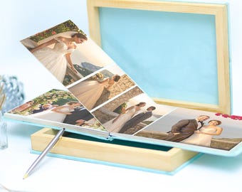 May include: A light blue wedding photo album with a wooden frame and a pen. The album is open, displaying several wedding photos. The album is approximately 12 inches long and 9 inches wide.