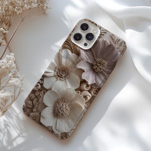 May include: A phone case featuring a 3D floral design in shades of beige and taupe. The case has a textured surface with three prominent flower shapes and intricate detailing. The camera lenses are visible at the top.