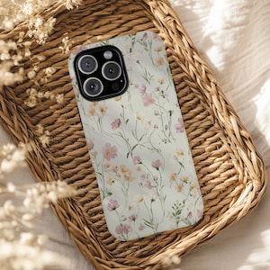 Pode incluir: Capa de telemóvel com padrão floral, com flores rosa e amarelas sobre um fundo claro. A capa está numa cesta de vime. O design floral evoca a natureza e a suavidade.