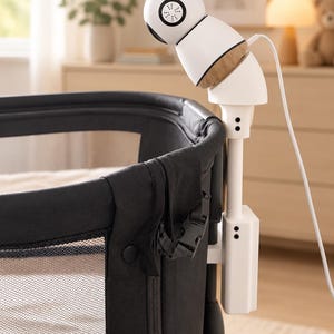 Peut inclure: Une caméra de surveillance pour bébé blanche et en bois clair fixée sur un berceau en maille noire. La caméra possède une tête sphérique avec un objectif noir et un câble d'alimentation blanc. Le bras de fixation blanc est fixé au cadre.