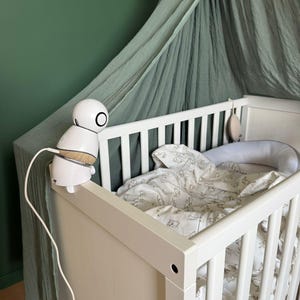 Op de afbeelding: Een witte babyfoon met een ronde kop en een houten basis is gemonteerd op een witte wieg. De wieg heeft een patroon deken en een bumper. Een groene luifel hangt boven de wieg.