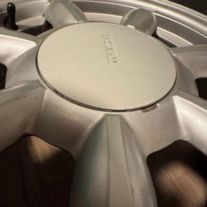 Puede incluir: Primer plano de una llanta de coche plateada con un neumático. La llanta tiene un tapacubos central con el logotipo "MAZDA" en relieve. El diseño presenta múltiples radios que se extienden desde el centro hasta el borde exterior.