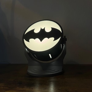 May include: A gray Batman-themed night light. The light features the iconic bat symbol in black against a white background. The light is mounted on a gray base and is angled upwards.