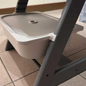 May include: A gray and white high chair with a tray and footrest. The chair has a rectangular tray and a footrest. The chair is made of plastic and metal. The chair is designed for infants and toddlers.