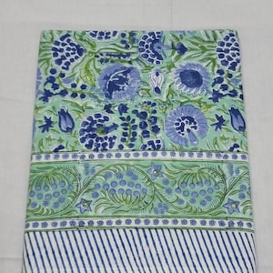 Puede incluir: Textil rectangular plegado con un estampado floral en tonos azules y verdes. El diseño incluye grandes flores estilizadas, hojas y un borde con rayas. La tela parece ser de algodón o un material similar.