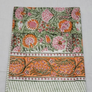 Op de afbeelding: Een gevouwen textiel met een bloemenpatroon. Het ontwerp heeft grote roze en oranje bloemen op een lichtgroene achtergrond. Een rand met oranje bloemelementen en een gestreepte rand maken het ontwerp compleet.